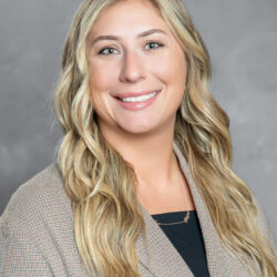 A woman with long, wavy blonde hair smiles at the camera, dressed in a checked blazer over a black top. The background is plain gray, and the image represents the Care Initiative brand.