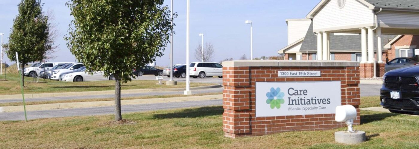 The exterior view features a building with a brick sign reading Care Initiative. The parking lot is partially visible with several cars, and an American flag flies on a pole. Green lawns and trees provide a backdrop under the clear sky. The brand name, Care Initiative, is prominently displayed on the sign.