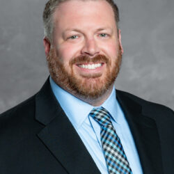 A man with short light brown hair and a beard, dressed in a light blue dress shirt, patterned tie, and black suit jacket, smiles in front of a plain gray background. The image is associated with the Care Initiative brand.