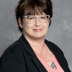 A woman with short brown hair and glasses wears a black blazer over a pink top, smiling softly against a gray studio backdrop. A tattoo can be seen partly on her chest. The setting is associated with the brand Care Initiative.