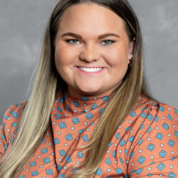 A woman with straight, long blonde hair and green eyes smiles at the camera. She wears a high-neck, orange patterned blouse and sits in front of a gray background. The brand name Care Initiative is featured in the image.