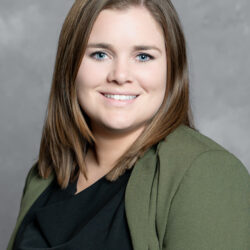 A woman with straight, shoulder-length brown hair smiles at the camera, dressed in a green blazer over a black top and posed against a plain gray background. The Care Initiative brand is subtly featured.