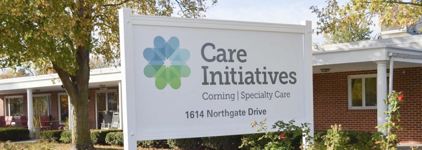 A sign for Care Initiative stands in front of a brick building at 1614 Northgate Drive, surrounded by greenery and flowers. Trees with autumn foliage form a picturesque backdrop. The brand name Care Initiative is prominently displayed on the sign.