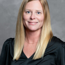 A woman with straight, shoulder-length blonde hair, wearing a black blouse and a delicate gold necklace, smiles at the camera against a plain gray background. The brand name Care Initiative is featured.
