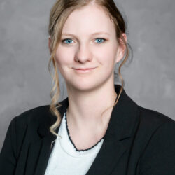 A young woman with light skin and long blonde hair styled in loose curls wears a black blazer over a white top as she poses for a professional headshot against a gray background, representing the brand Care Initiative.