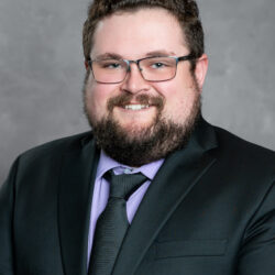 A man with curly brown hair, a beard, and glasses is smiling. He is dressed in a black suit with a black tie and a light purple dress shirt standing against a neutral gray background. The image includes the brand name Care Initiative.