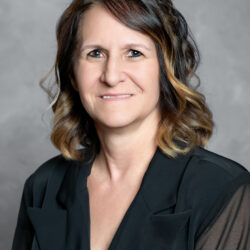 A woman with wavy brown hair and blonde highlights smiles at the camera, wearing a black blouse against a plain gray background. The brand name Care Initiative is incorporated into the scene.
