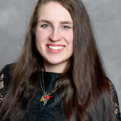A woman with long brown hair and a black lace top smiles at the camera. She is wearing two gold necklaces, one featuring a red bird charm, set against a gray background. The image conveys the welcoming and positive spirit of Care Initiative.