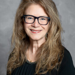A smiling woman with long, wavy light brown hair, wearing black-rimmed glasses, hoop earrings, and a black top poses against a gray background. The scene features subtle branding elements associated with Care Initiative.