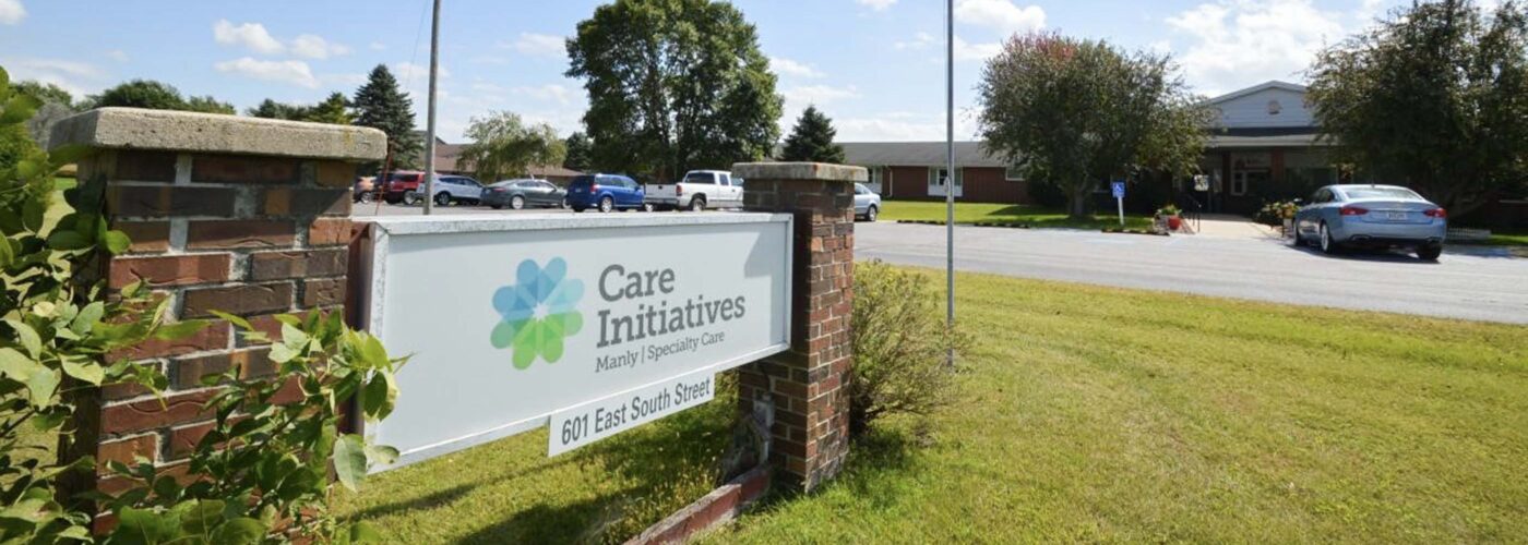 A Care Initiative sign stands prominently in front of a healthcare facility. Nearby, the American flag flutters on a flagpole. The scene features a grassy area, driveway, and cars parked under a partly cloudy sky. The brand name Care Initiative is clearly visible on the sign.