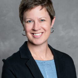 A woman with short brown hair, wearing hoop earrings, a light blue top, and a dark blazer smiles at the camera against a plain gray background, representing Care Initiative.