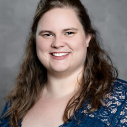 A woman with long brown hair smiles, wearing a dark blue lace top, posed in front of a neutral gray background. Care Initiative.