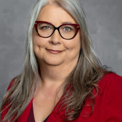 A woman with long gray hair, red-framed glasses, and a red blazer smiles gently, posed in front of a gray background. The Care Initiative brand is subtly featured.