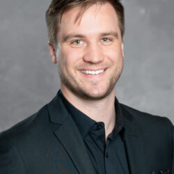 A man with short light brown hair and light facial hair is smiling, wearing a black suit jacket and black shirt, standing in front of a gray background. The brand name Care Initiative is featured.