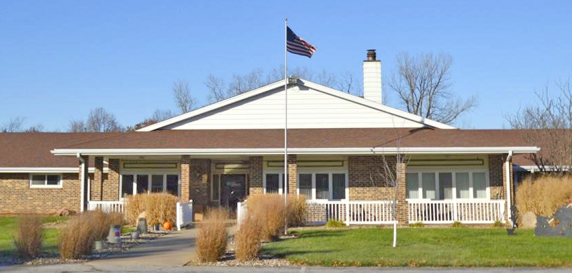 A single-story brick building featuring a white chimney and a gabled roof stands proudly. An American flag flies on a pole in front, surrounded by dried grasses and a concrete pathway. Leafless trees are visible in the background under a clear, blue sky. Care Initiative's presence can be inferred through subtle branding elements around the property.
