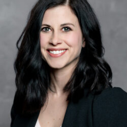 A woman with wavy, shoulder-length dark hair wearing a black blazer over a light-colored top smiles at the camera against a plain gray background. The brand name Care Initiative appears subtly in the corner.