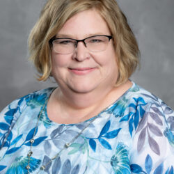 A woman with light hair and glasses smiles at the camera, wearing a blue and white floral top and a silver necklace against a gray background. The Care Initiative brand is associated with her portrait.
