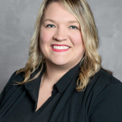 A woman with blonde, wavy hair wearing a black top and blazer smiles at the camera against a plain gray background, representing Care Initiative.