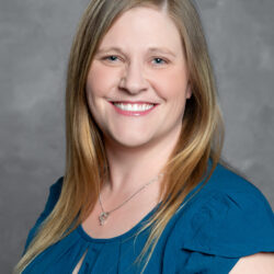 Smiling woman with straight blonde hair wearing a blue blouse and a heart-shaped necklace posing in front of a plain gray background Care Initiative.