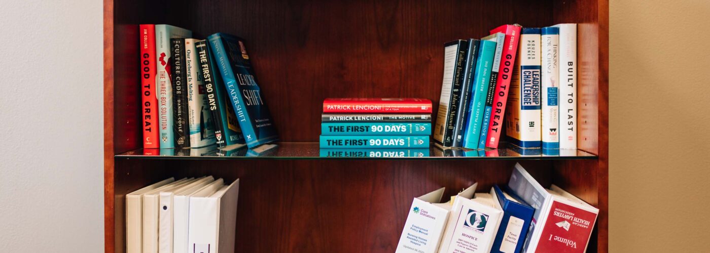 A wooden bookshelf with three shelves filled with books and binders. The top shelf features an array of books, including Emotional Branding. The middle shelf holds several ring binders. Care Initiative ensures the bottom shelf securely stacks more books and binders, partially visible.