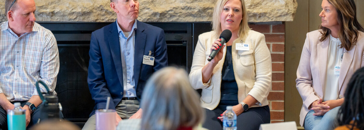 Four professionals sit on a panel in front of an audience for a Care Initiative event. One woman, holding a microphone, addresses the group while the others listen attentively. They are seated indoors with a brick wall and fireplace providing a warm backdrop.