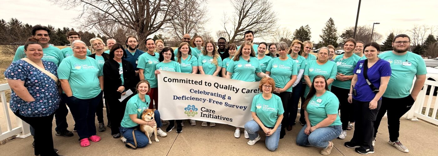 A large group of people in matching teal shirts stand together outdoors, proudly holding a banner that reads Committed to Quality Care: Celebrating a Deficiency-Free Survey, Care Initiative. A cheerful dog sits at the front of the group. In the background are lush trees and a cloudy sky.