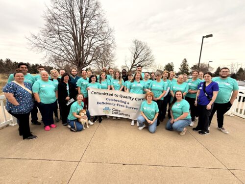 A large group of people in matching teal shirts stand together outdoors, proudly holding a banner that reads Committed to Quality Care: Celebrating a Deficiency-Free Survey, Care Initiative. A cheerful dog sits at the front of the group. In the background are lush trees and a cloudy sky.