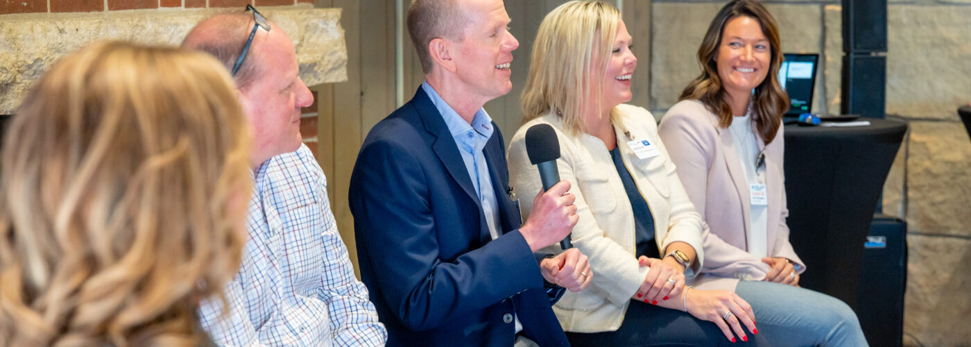 Four people sit next to each other, smiling and engaged in conversation. The man in the center holds a microphone, suggesting a panel discussion for Care Initiative. A woman in the foreground faces the group. In the background, there is a screen and speaker positioned behind them.