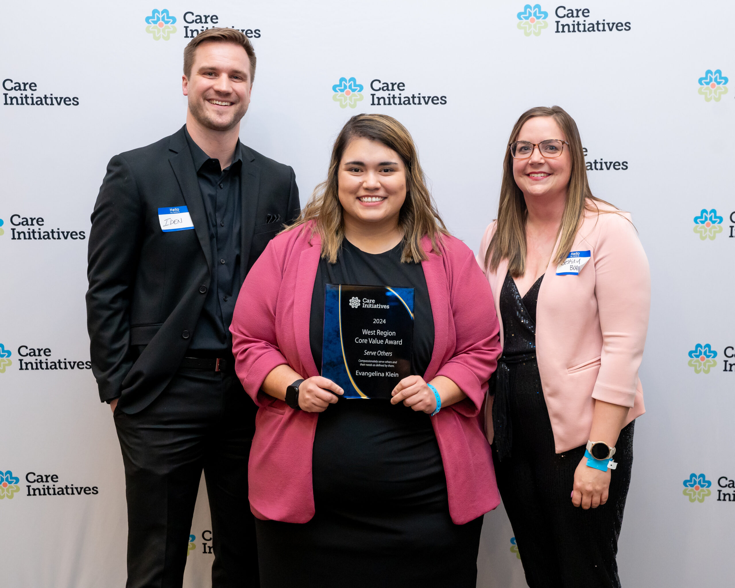 Three people stand smiling in front of a Care Initiatives backdrop. The person in the center holds an award plaque. All are dressed in business attire and wearing name tags, representing Care Initiatives.