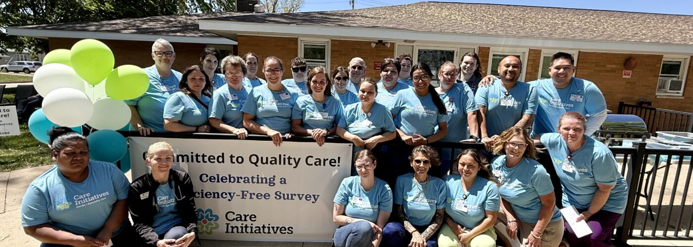 A large group of people in matching light blue shirts stand together outside a building, smiling proudly in front of a colorful banner that reads Committed to Quality Care! with festive balloons on the side. The brand name Care Initiative is prominently displayed, emphasizing their dedication and team spirit.