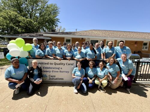 A large group of people in matching light blue shirts stand together outside a building, smiling proudly in front of a colorful banner that reads Committed to Quality Care! with festive balloons on the side. The brand name Care Initiative is prominently displayed, emphasizing their dedication and team spirit.