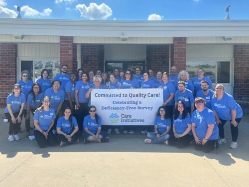 A large group of people in blue shirts stand outside a building, smiling and holding a banner that reads: Committed to Quality Care! Celebrating a Deficiency-Free Survey. The Care Initiatives brand is prominently displayed on the banner.