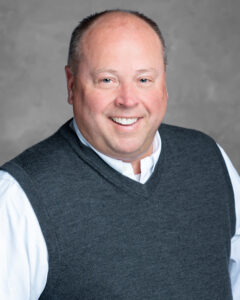A middle-aged man with short light hair in a white shirt and dark gray sweater vest smiles at the camera, Care Initiative backdrop.