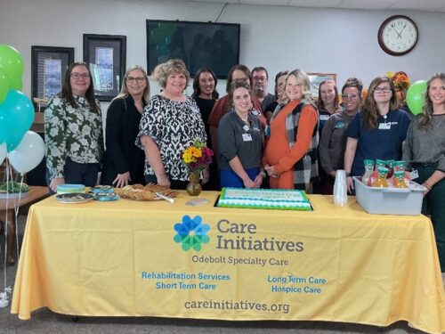 People smile behind a yellow Care Initiative table with cake and snacks, green and white balloons frame the festive indoor scene.