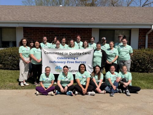 A group in light green Care Initiative shirts stand and sit outside a brick building in front of a banner about quality care.