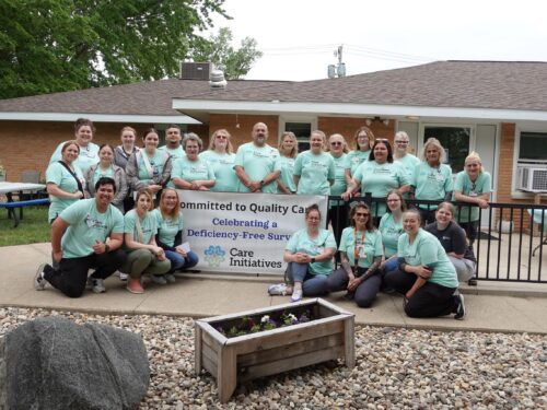 Team in light blue shirts smiles outside a building around a banner celebrating a deficiency-free survey for Care Initiative.