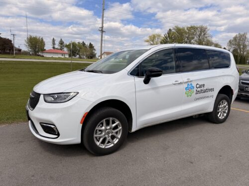 A white minivan featuring the Care Initiative logo and website is parked on a paved road beside grass, trees, and distant buildings.